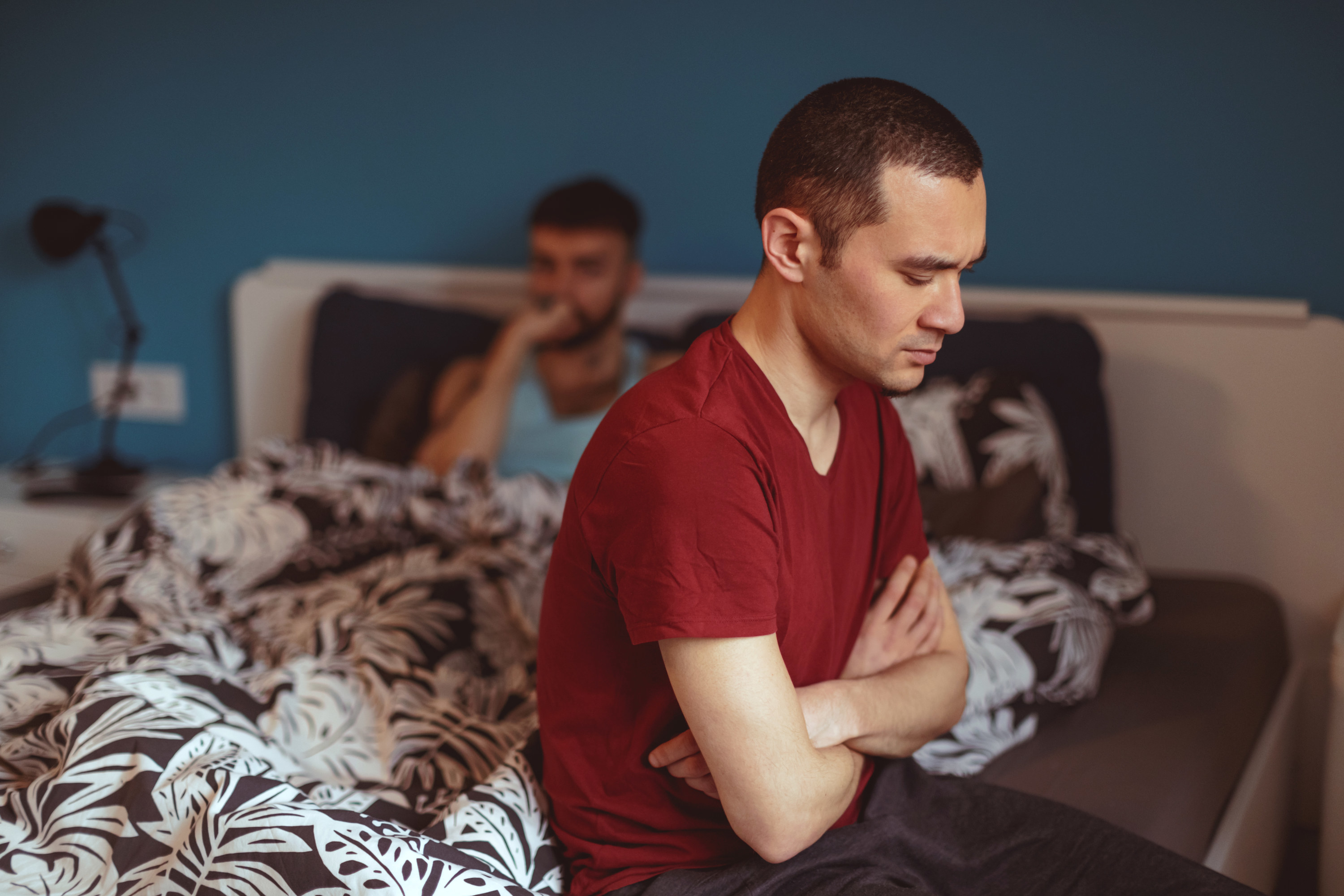 An upset man sits on the edge of the bed while his partner lies under the covers