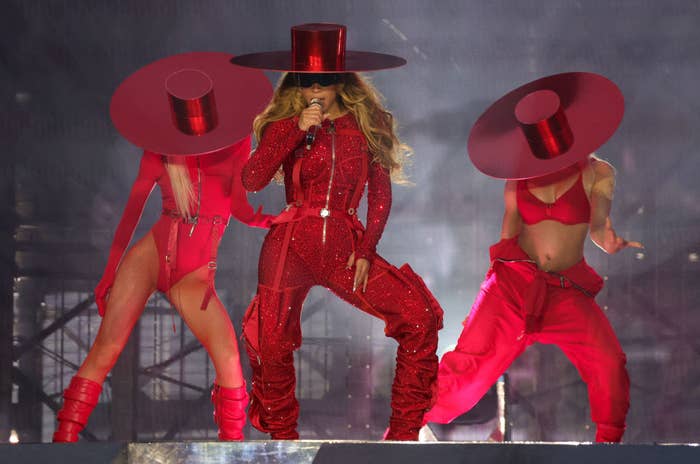 Beyoncé on stage wearing a sparkly jumpsuit with a matching wide-brimmed hat as two people dance behind her