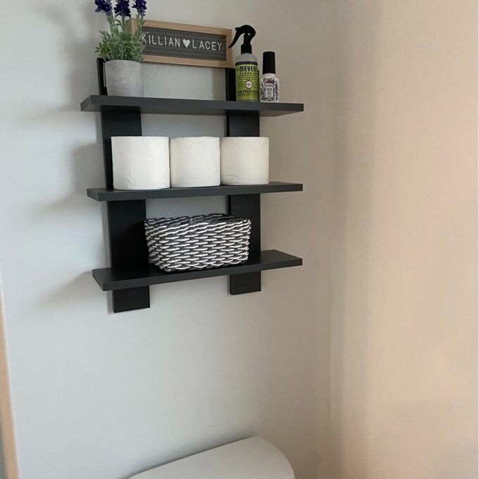 Black tiered storage shelf over toilet holding basket, toilet paper rolls, plant and toiletries