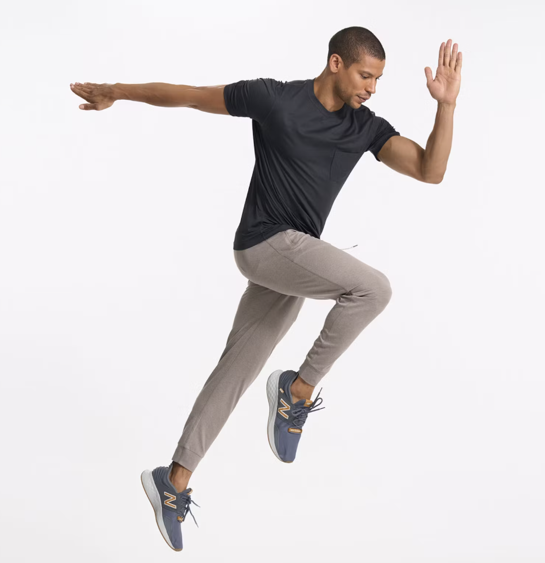 a man jumping in the air with his arms and legs extended wearing Vuori gray joggers