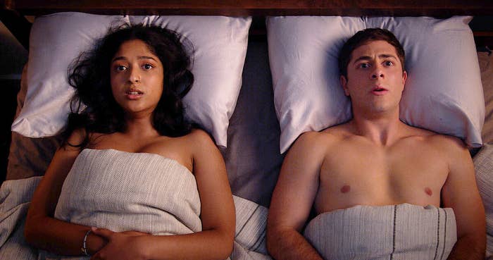 Devi and Ben lay naked and covered with a blanket on a bed with uncomfortable looks on their faces.