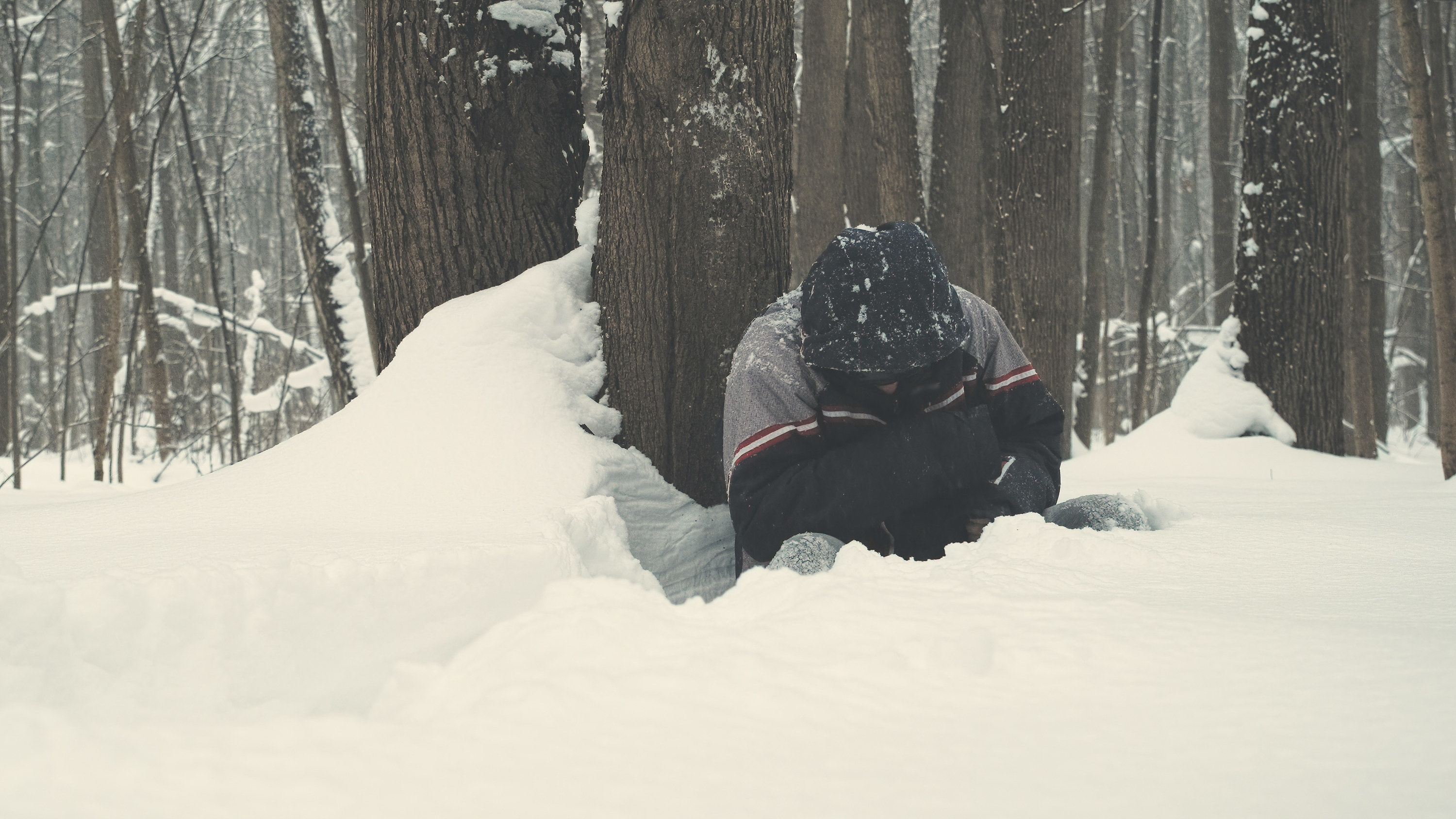 man wearing sunglasses sits waist deep in snow next to a tree and falls asleep in cold