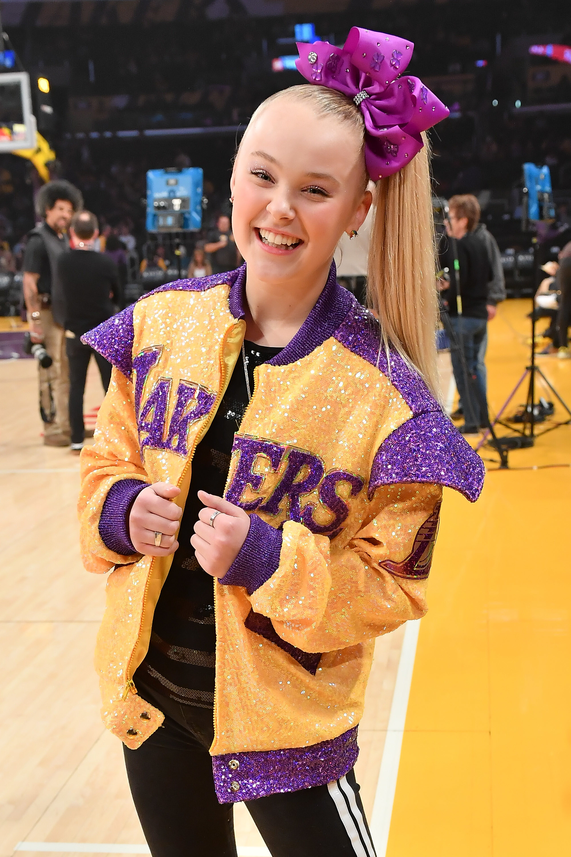 JoJo smiling on the basketball court wearing a sequined Lakers jacket and her signature side ponytail