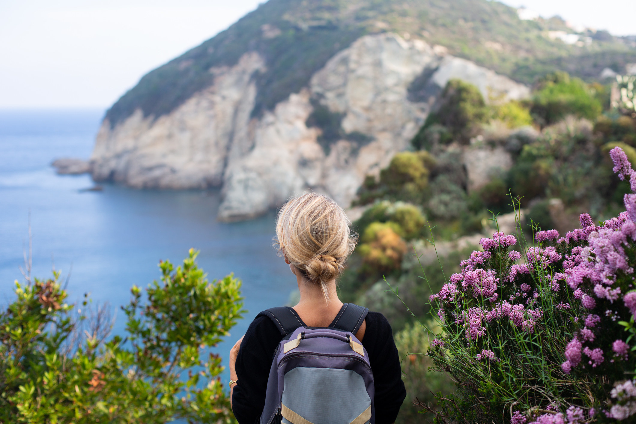 A woman with a backpack hiking on a footpath and looking at the rocky, verdant coastline