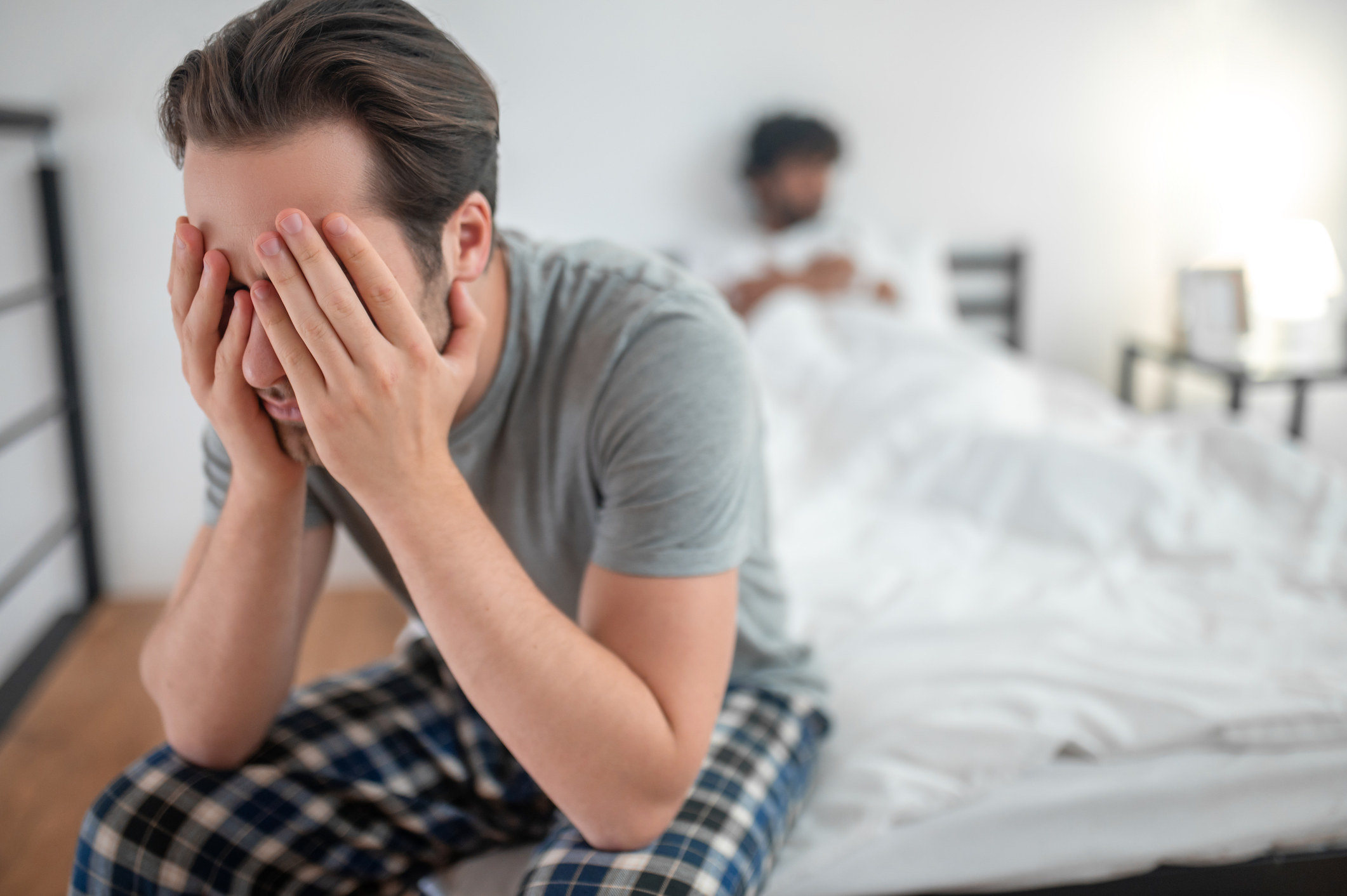 A man with his head in his hands on the edge of the bed with another man laying in the bed in the background