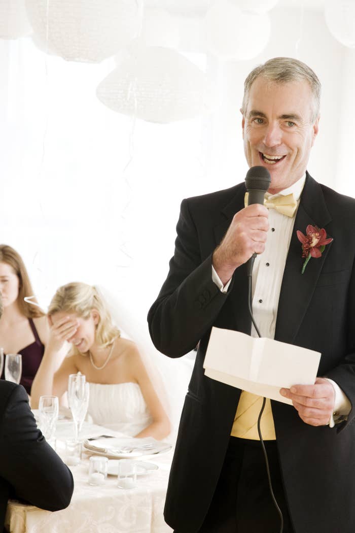 At a wedding, a man who is presumably father of the bride gives a speech while people laugh in the background