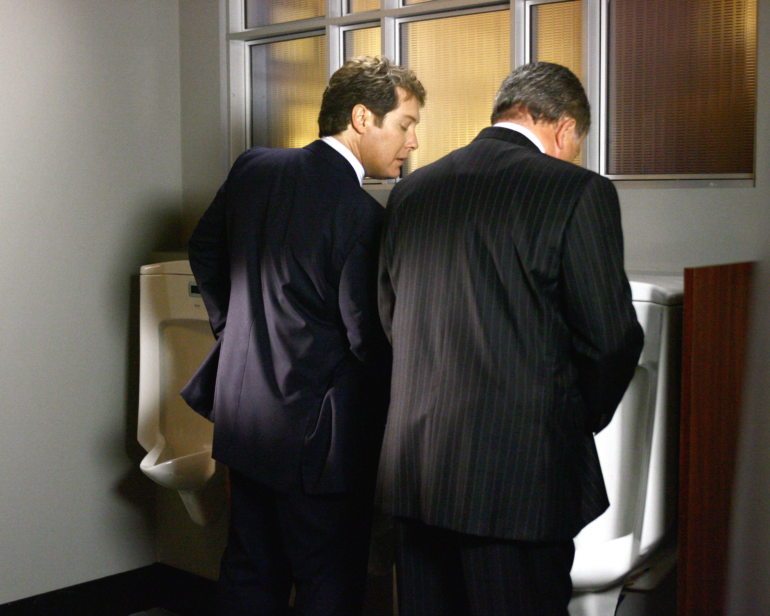 Two men standing at a urinal and one man is looking down at what the other man is doing