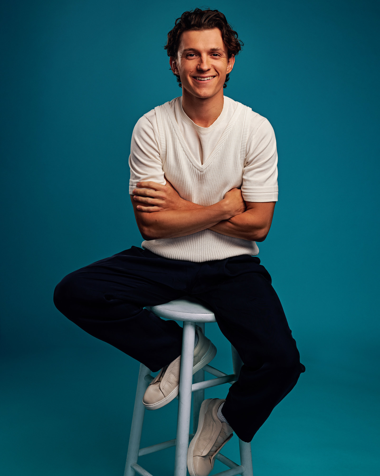 Tom Holland smiles with his arms crossed for a portrait as he sits on a stool