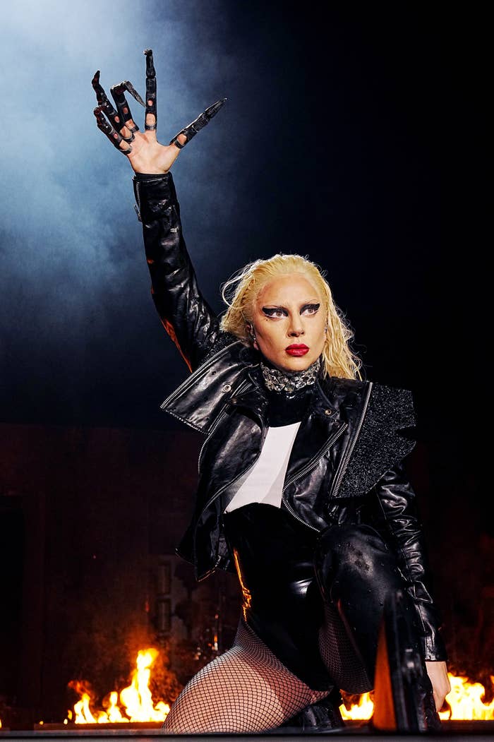 Lady Gaga raises an arm in the air as she performs with fire in the background. Each finger on her hand has its own individual glove