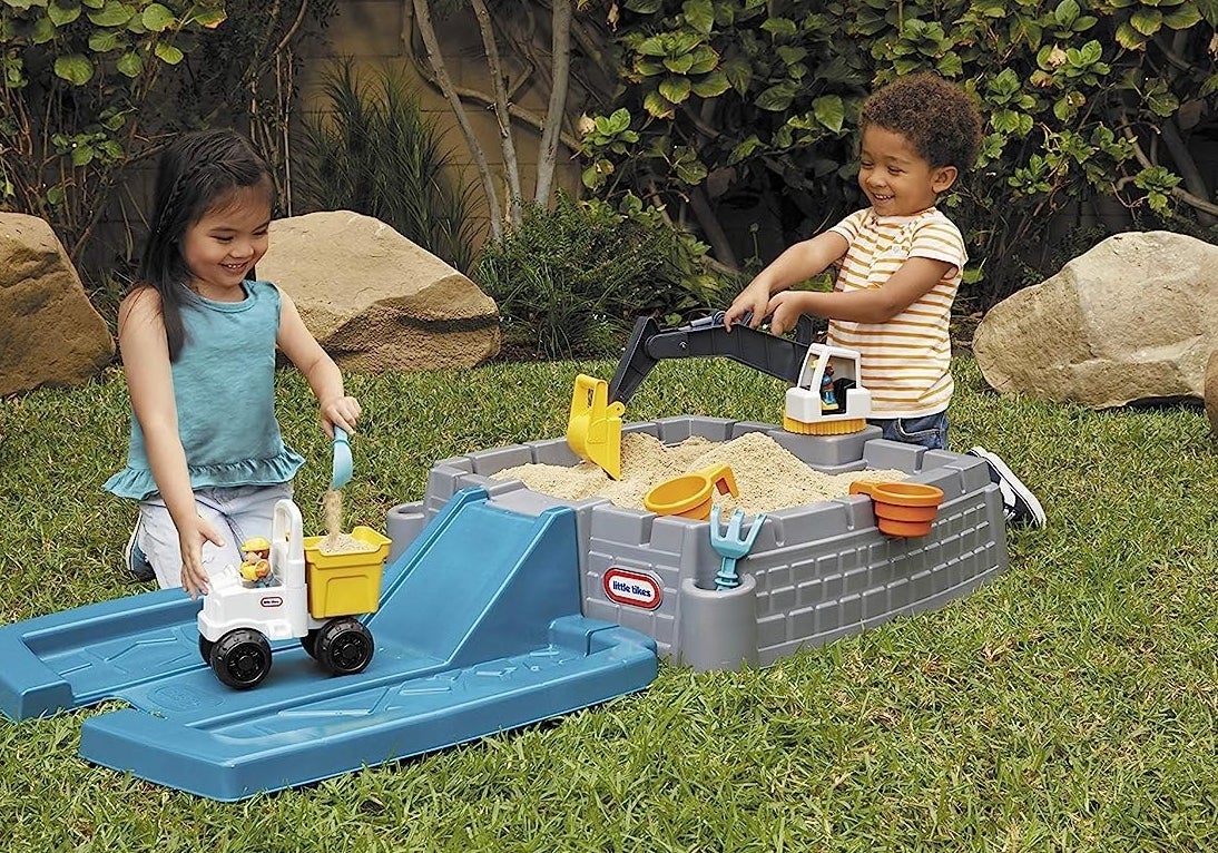 Two child models playing with blue and gray plastic sandbox with yellow digging toys