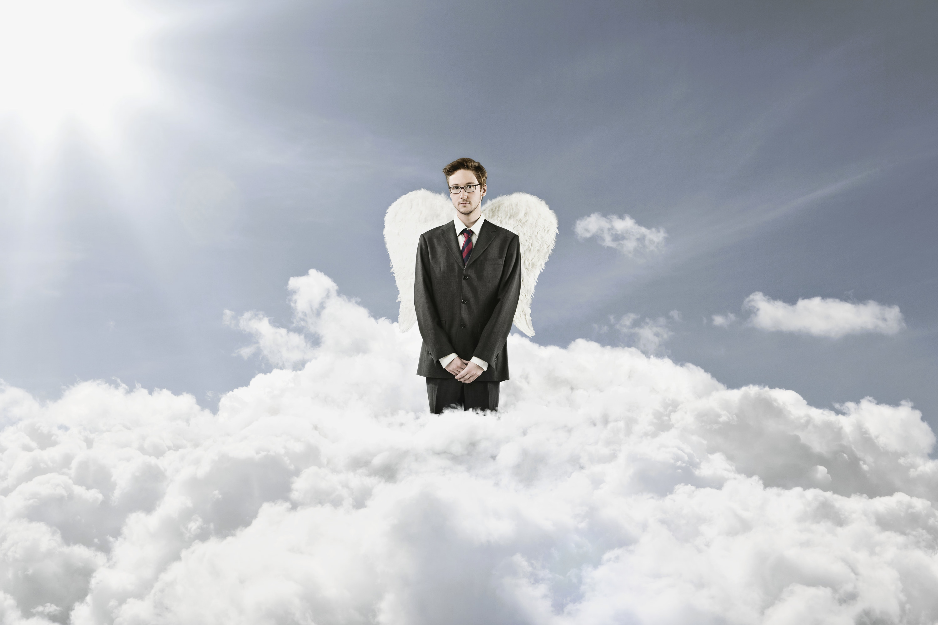A man in a business suit stands in the midst of clouds and has angel wings