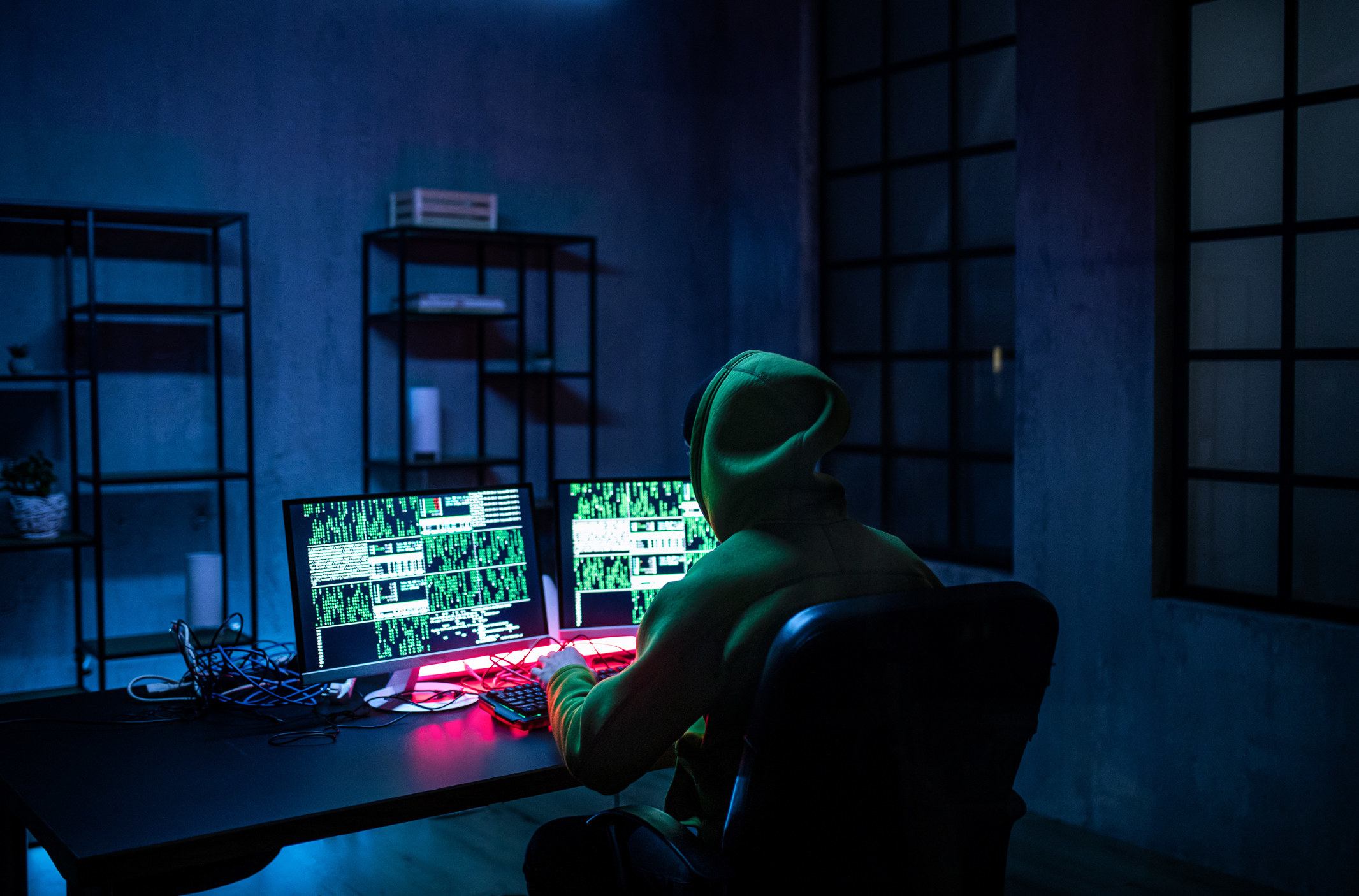 A computer hacker sits in a hoodie in a dark room