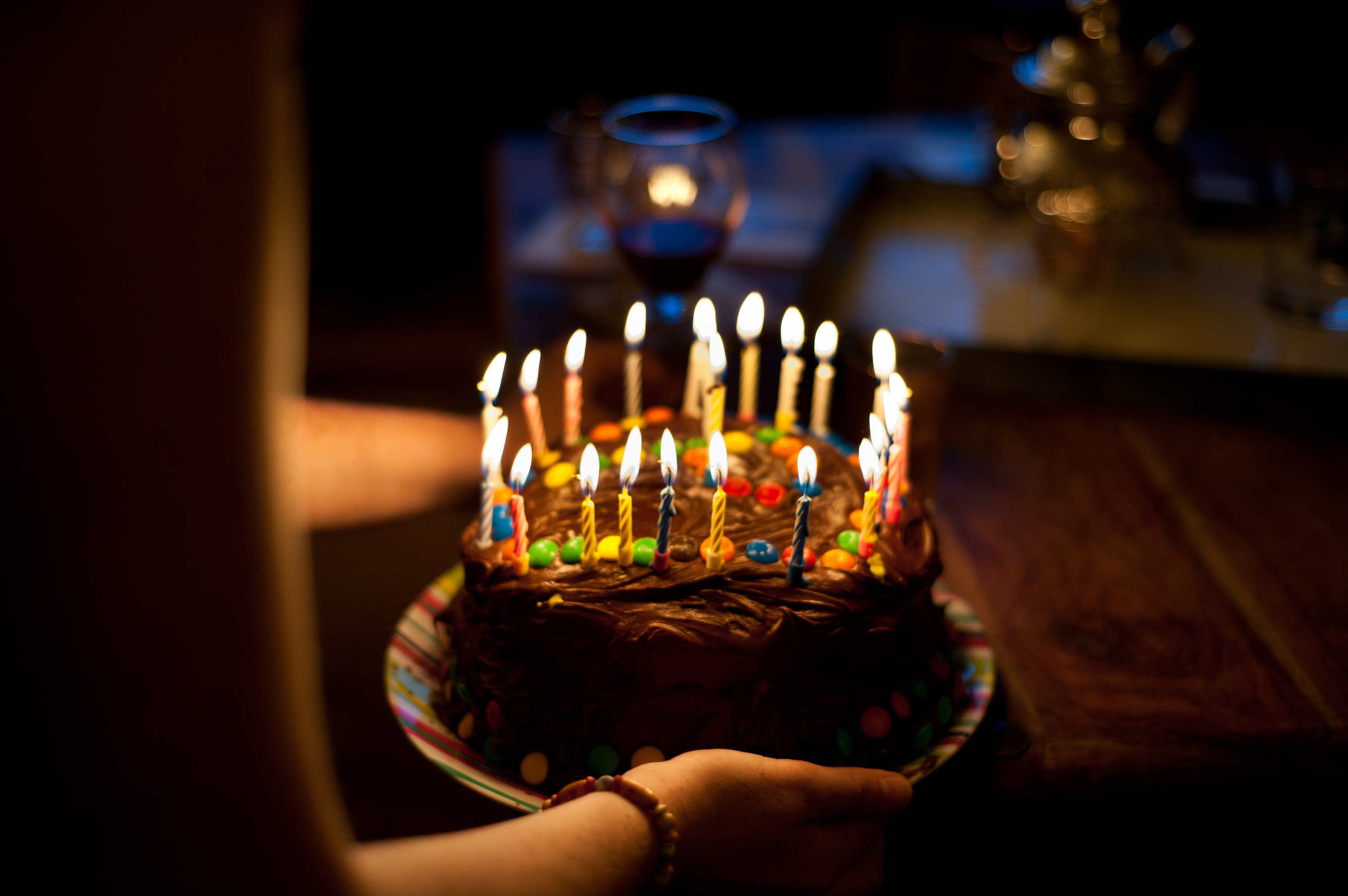 A person holds a chocolate frosting and M&amp;amp;M cake topped with lit candles at a birthday celebration