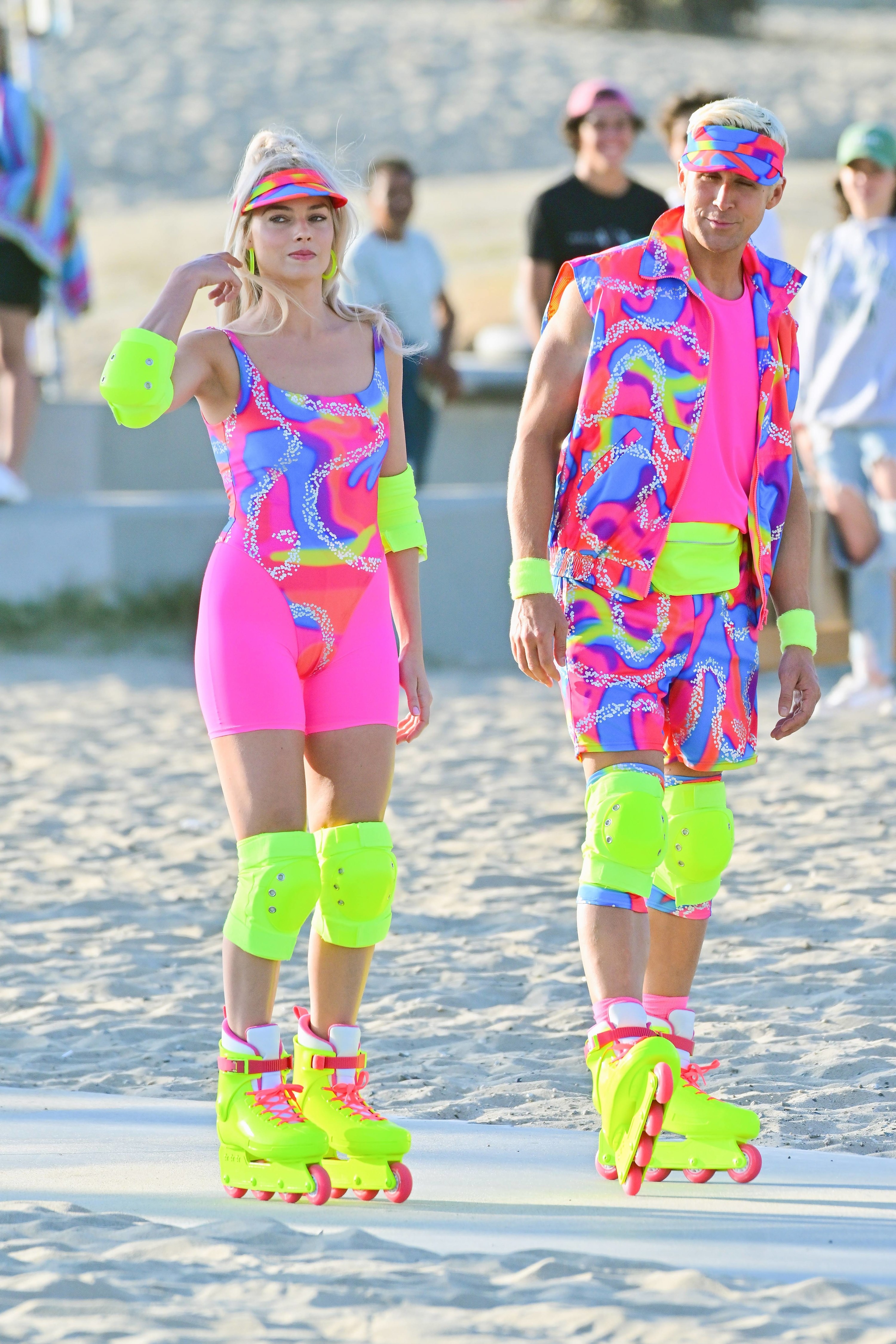 Margot and Ryan roller-skating in matching neon-colored outfits that include knee pads and visors