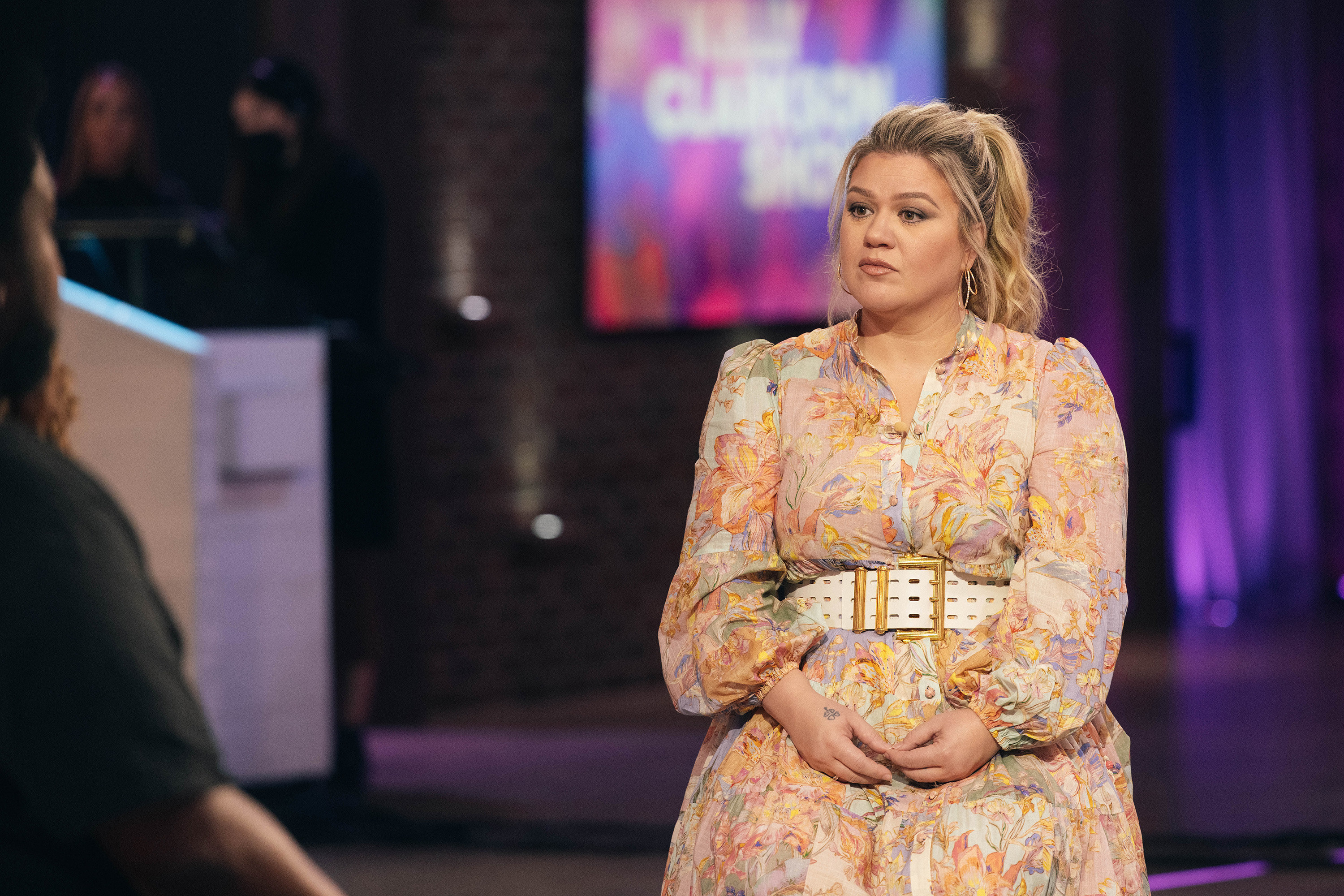 Kelly seated and wearing a long-sleeved, belted, floral-print dress and looking serious