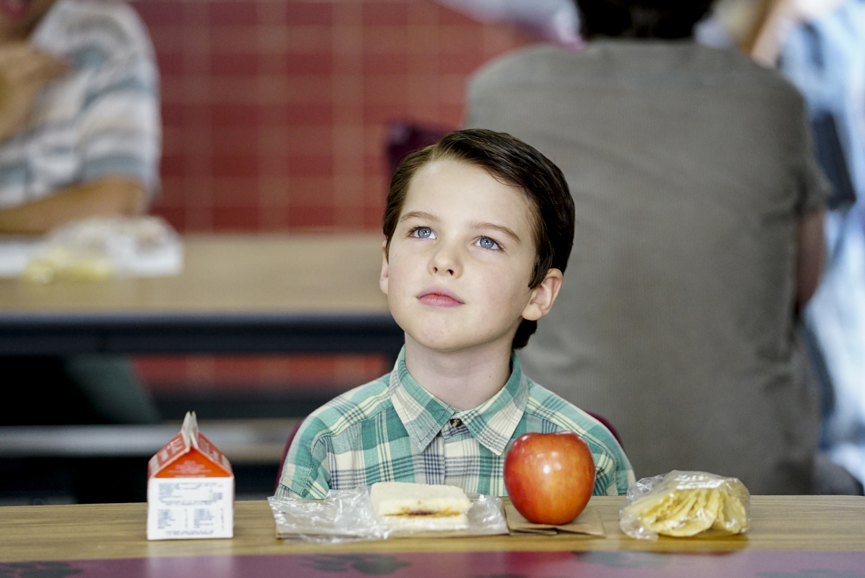 young sheldon at school lunch with an apple, sandwich, and milk in front of him