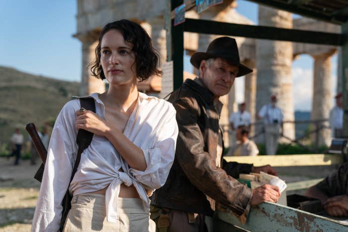 Indiana leans against a railing and looks at Helena who's carrying a backpack