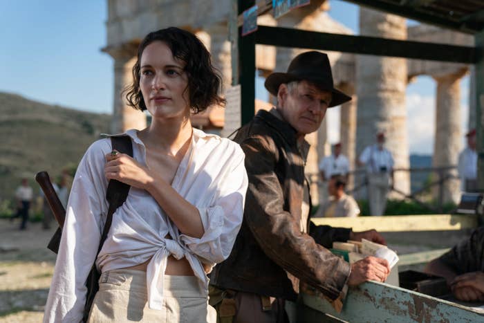 Indiana leans against a railing and looks at Helena who&#x27;s carrying a backpack