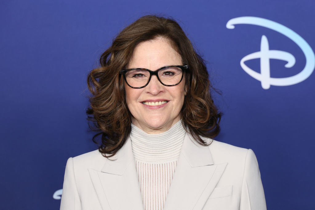 Close-up of a smiling Ally wearing glasses and a suit jacket and turtleneck