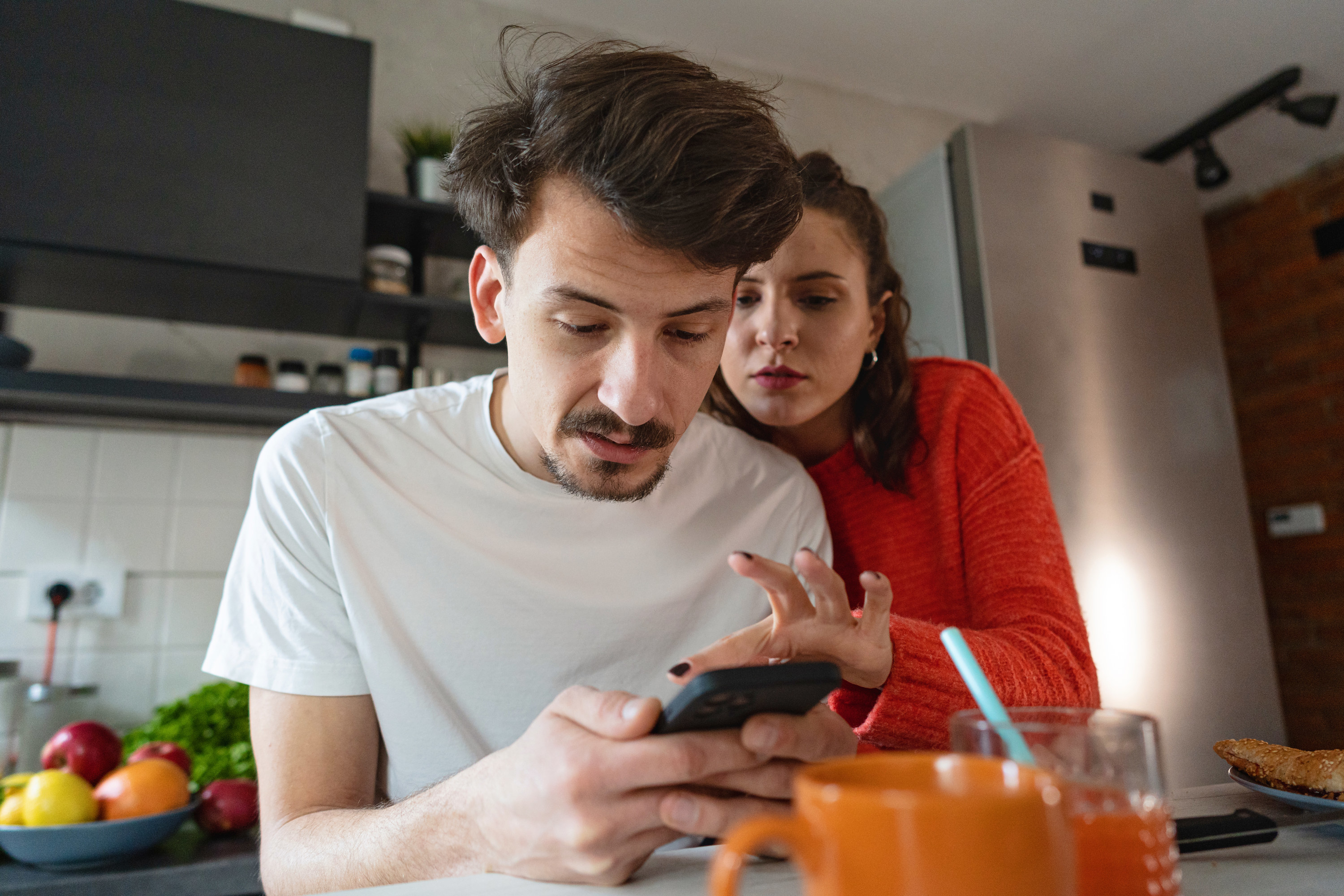 A woman looks through her boyfriend&#x27;s phone while the two stand in their kitchen