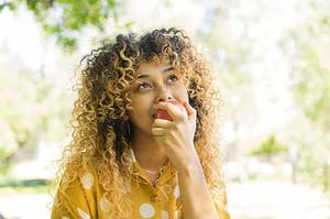 Mixed race woman with curly hair sitting on a park bench eating an apple looking into the sky 