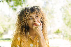 Mixed race woman with curly hair sitting on a park bench eating an apple looking into the sky 