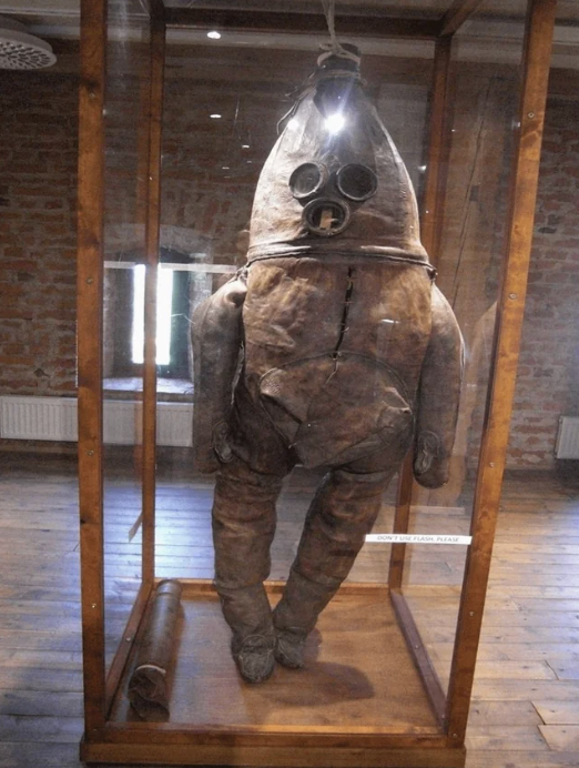 an old, massive brown diving suit, likely made of animal skin, in a glass case