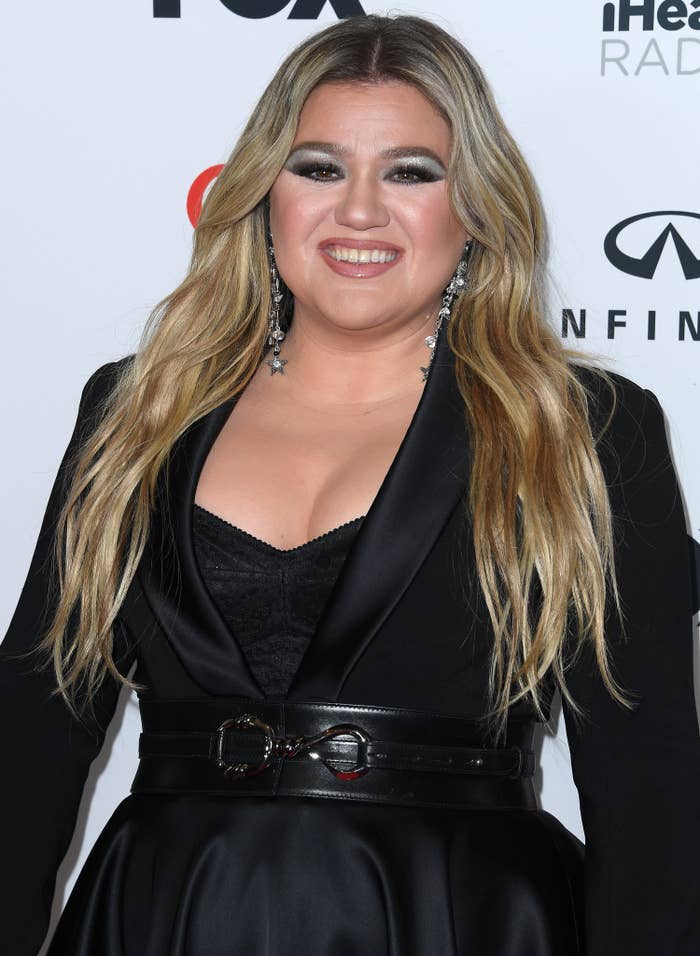A closeup of Kelly smiling at a red carpet event. She&#x27;s wearing a belted jacket and a bustier top underneath
