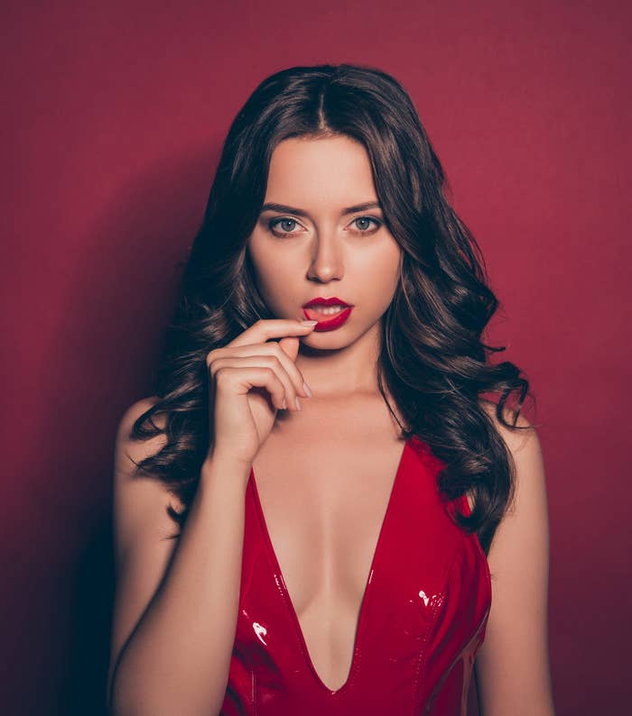 Woman with long hair and a deep-V sleeveless outfit touching her lower lip and staring into the camera