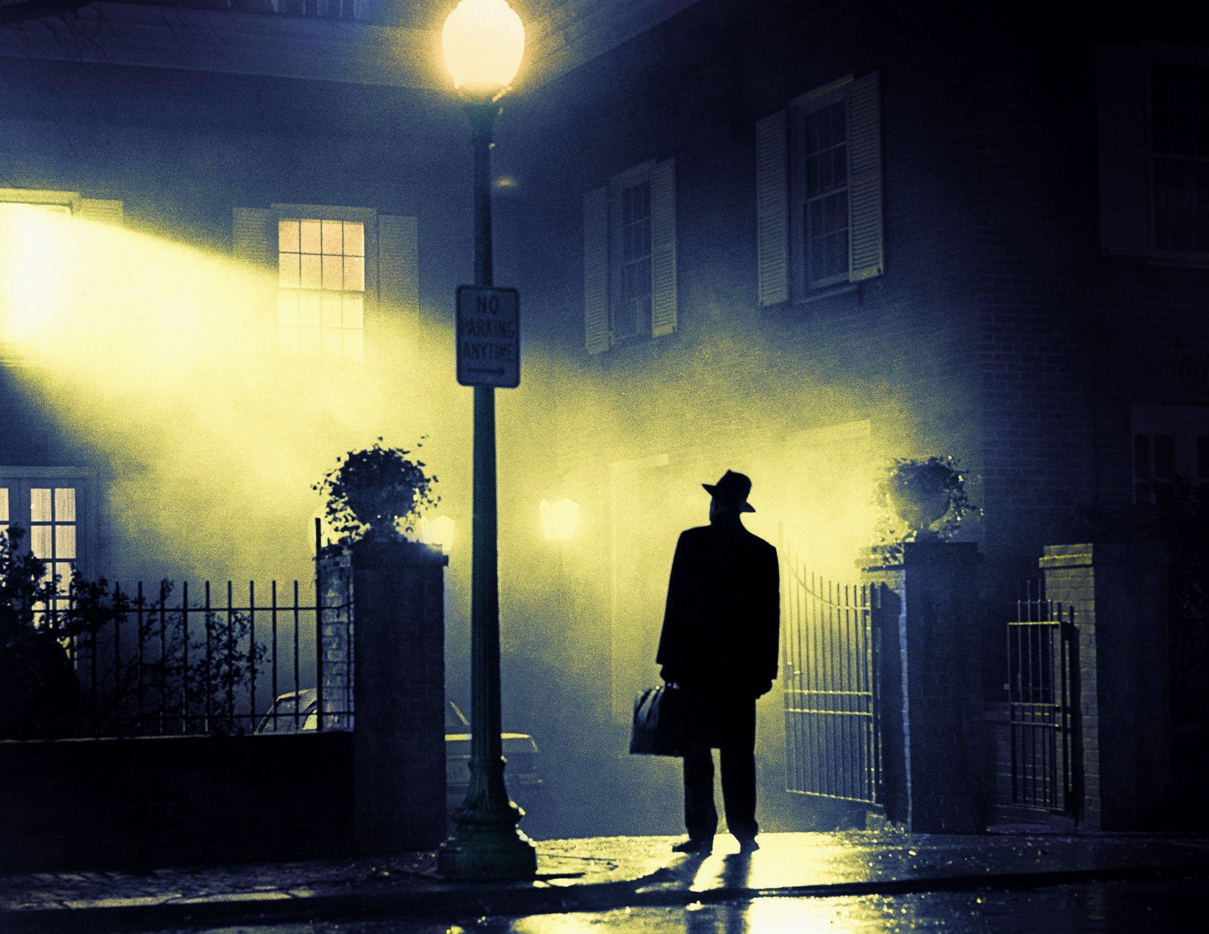 A man with a hat and briefcase basks in a light outside of a home at night