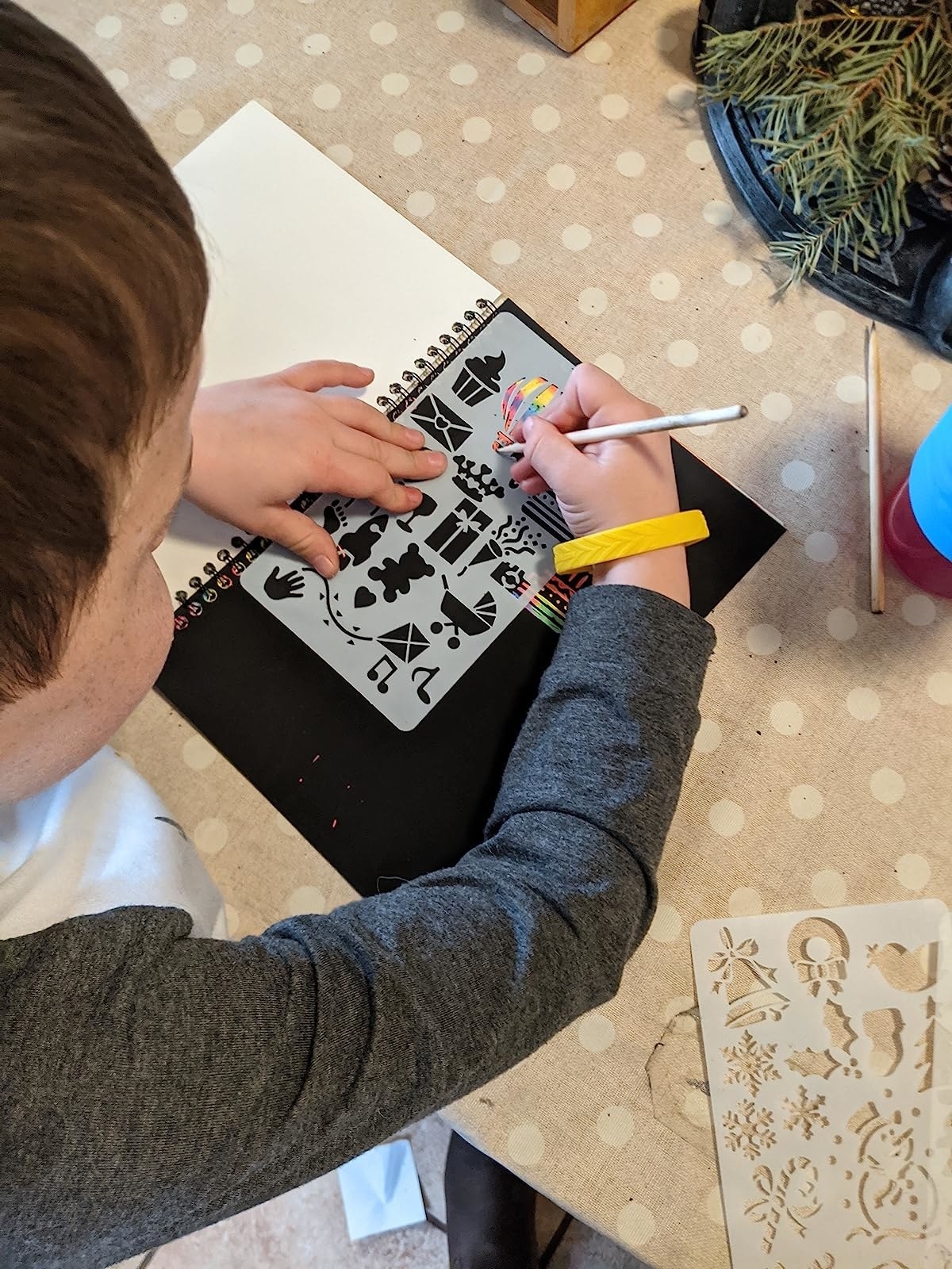 Reviewer&#x27;s young child using stencil to scratch rainbow picture on black page of the notebook