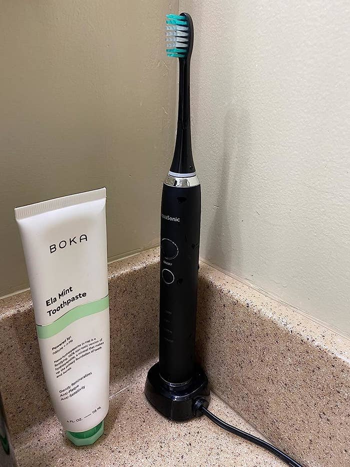the black toothbrush sitting on charging dock on reviewer&#x27;s bathroom counter next to mint toothpaste