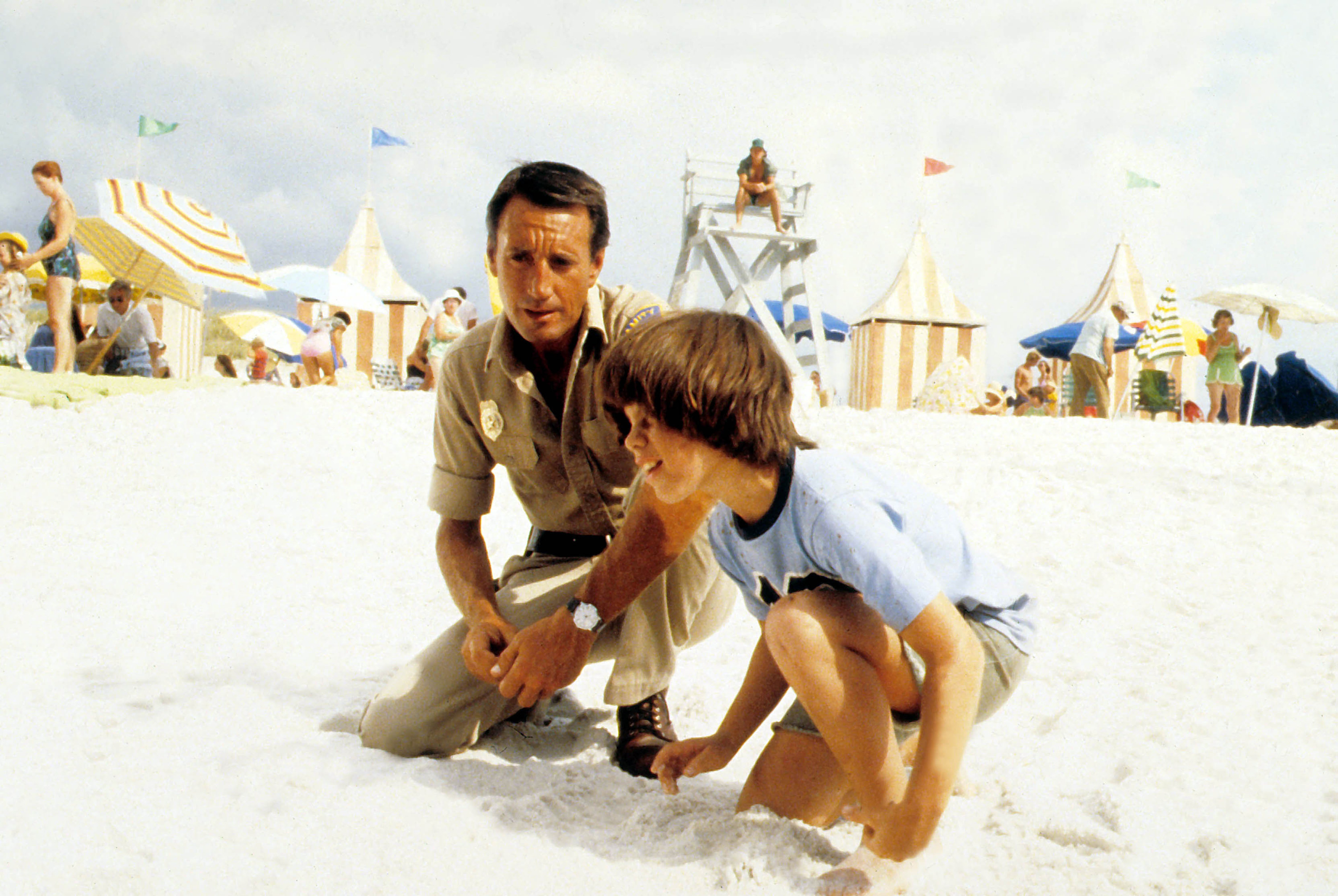 Roy Scheider kneels on a beach near a young boy