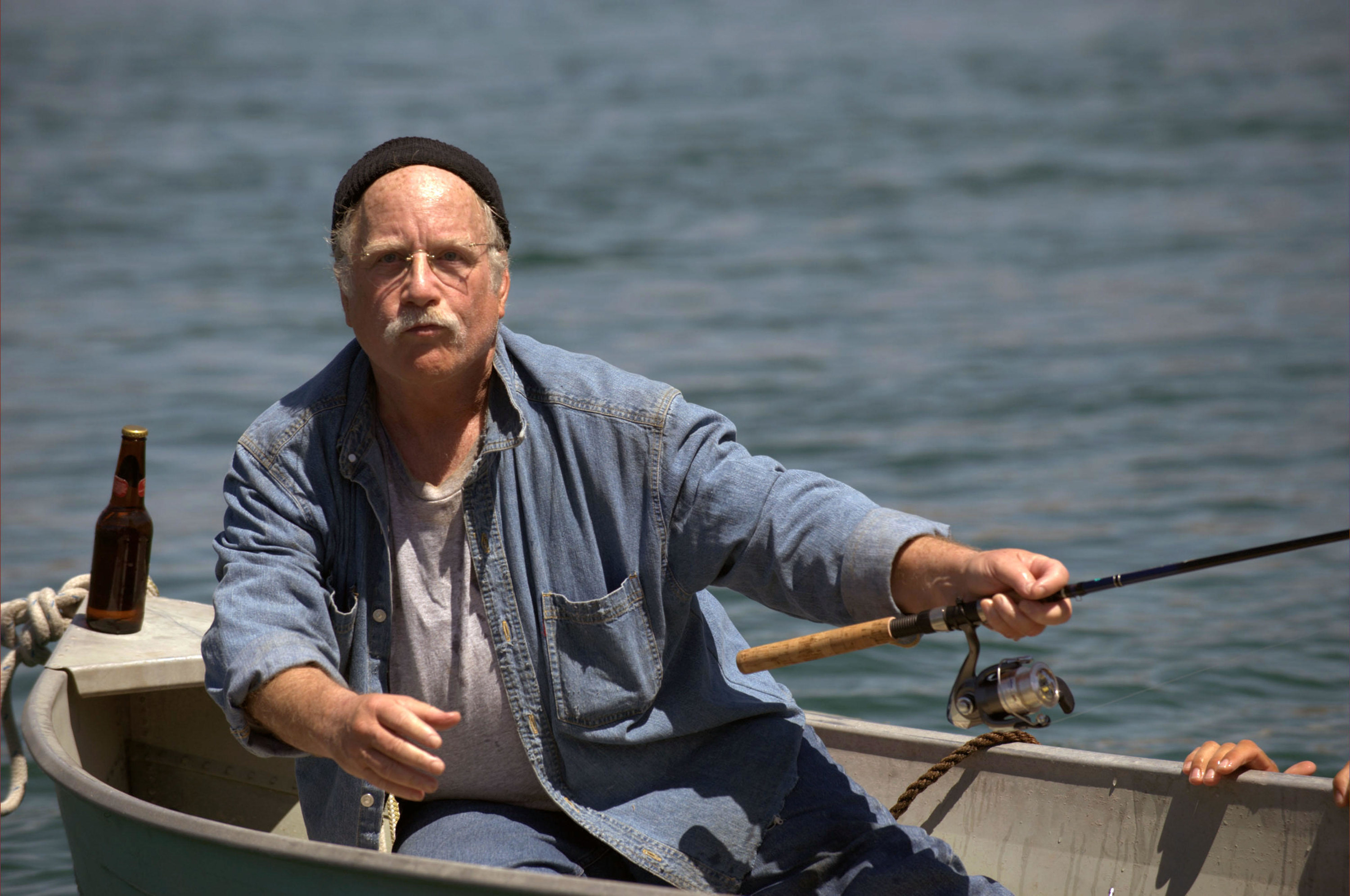 Richard Dreyfuss sits on a small boat with a bottle of beer and a fishing pole