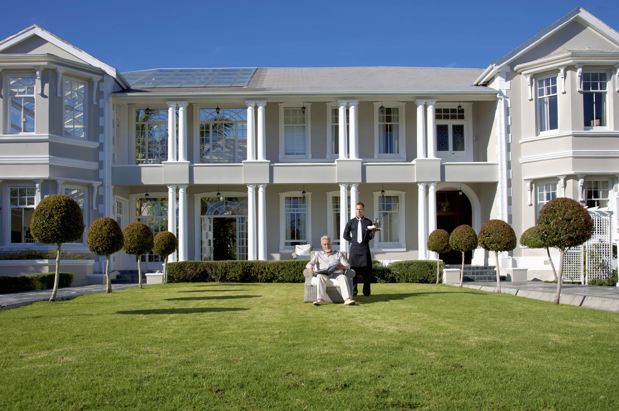 A butler bringing a tray to a man in his backyard outside of a large mansion