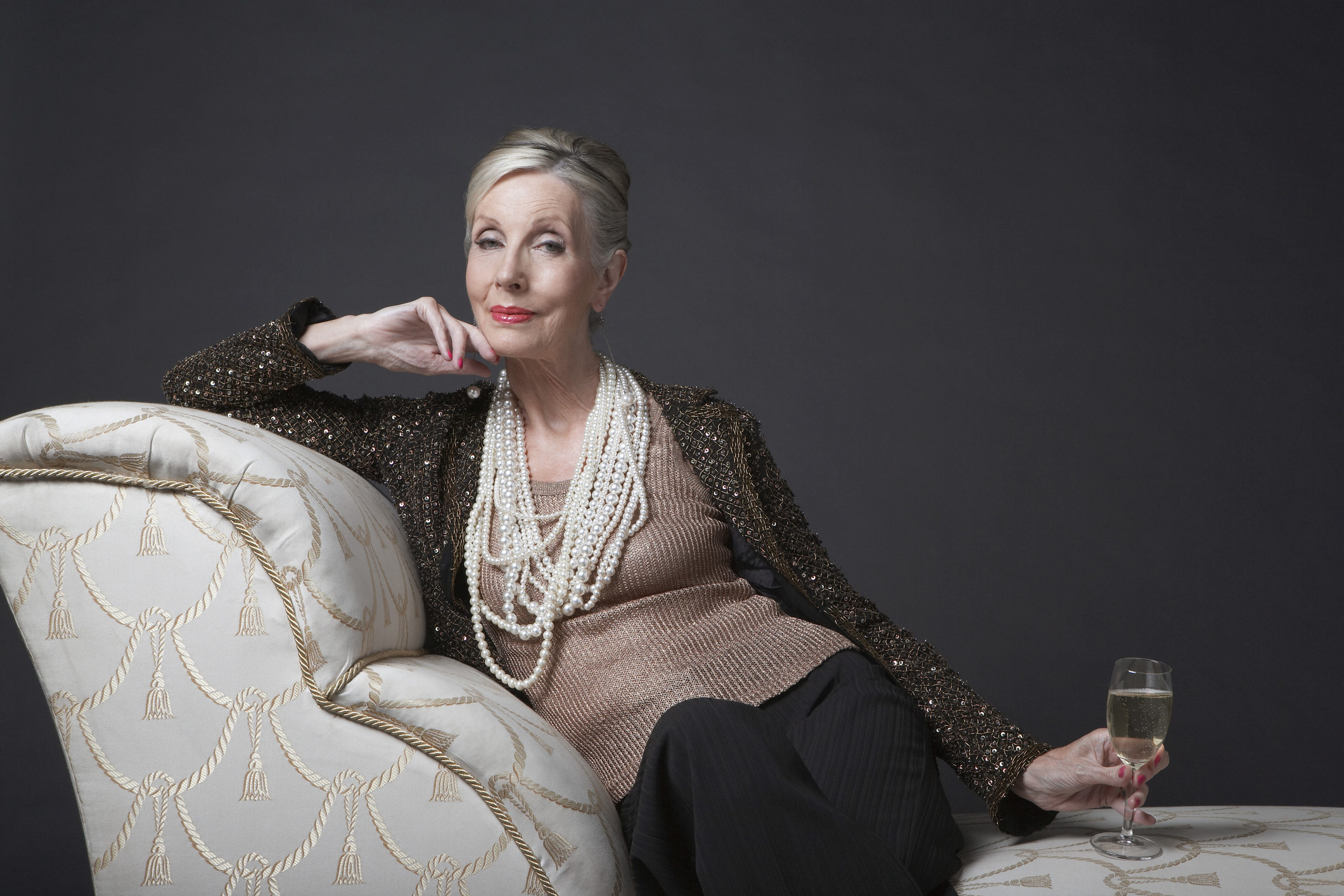 A woman sitting on a chaise lounge with a glass of champagne in her hand and pearls around her neck