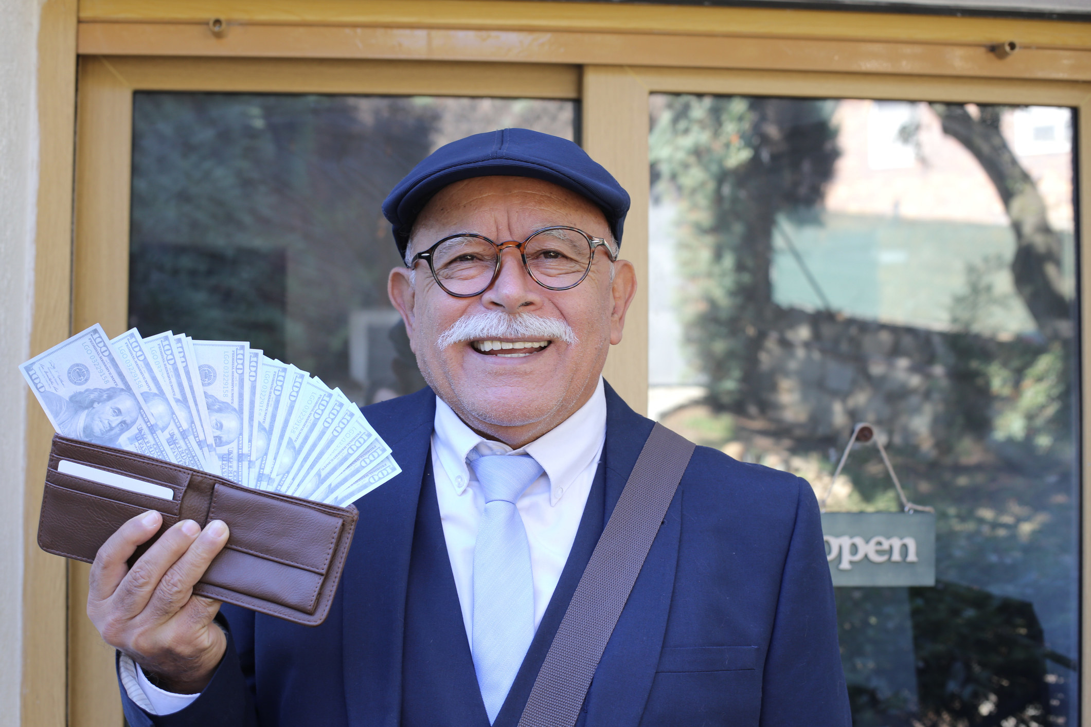 A man with a mustache and glasses holding up his wallet which has a lot of cash in it