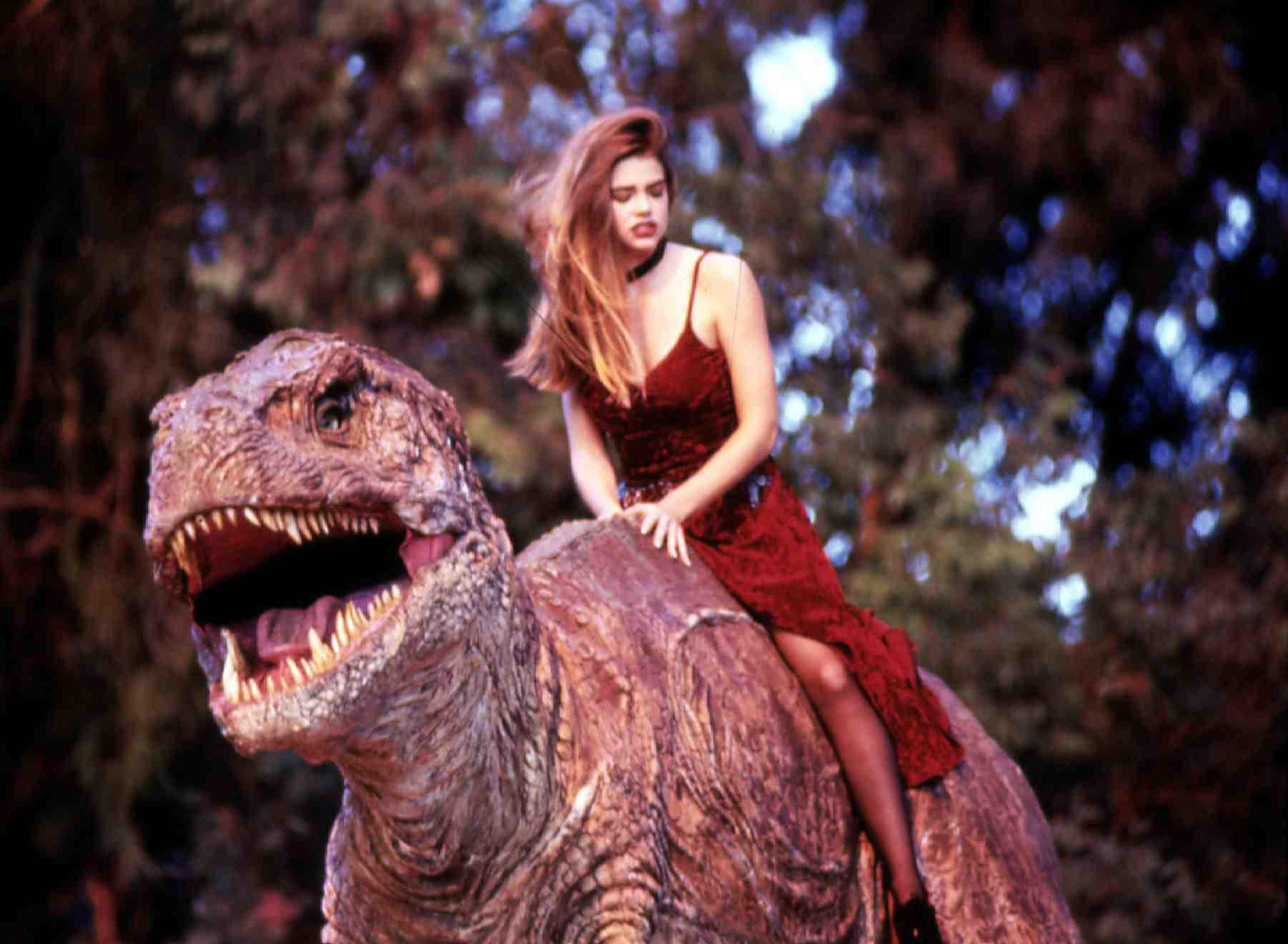 A young Denise Richards in a red dress rides a T-rex