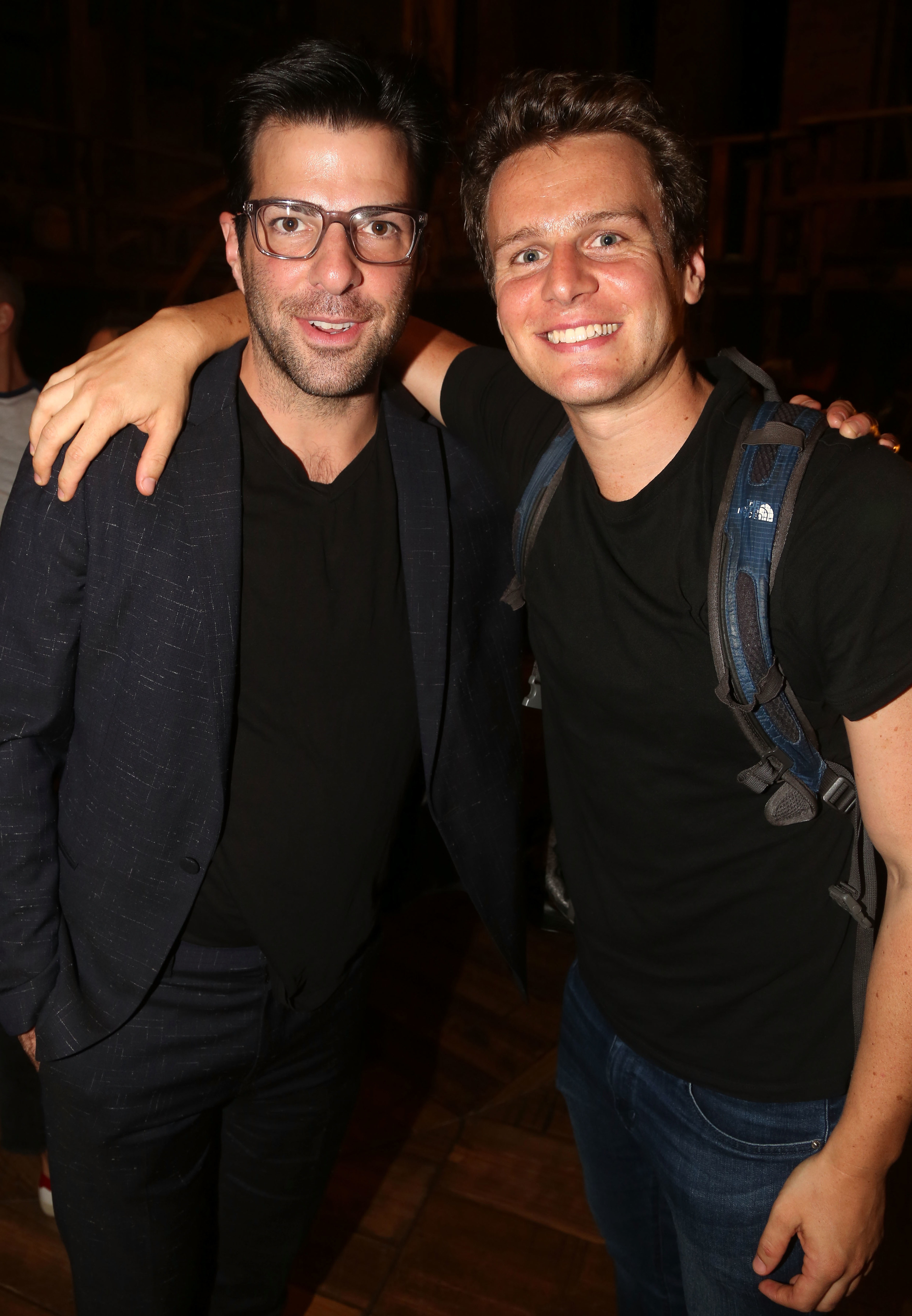 Zachary Quinto wearing a blazer and pants and Jonathan Groff wearing a plain dark t-shirt and jeans with their arms around each other