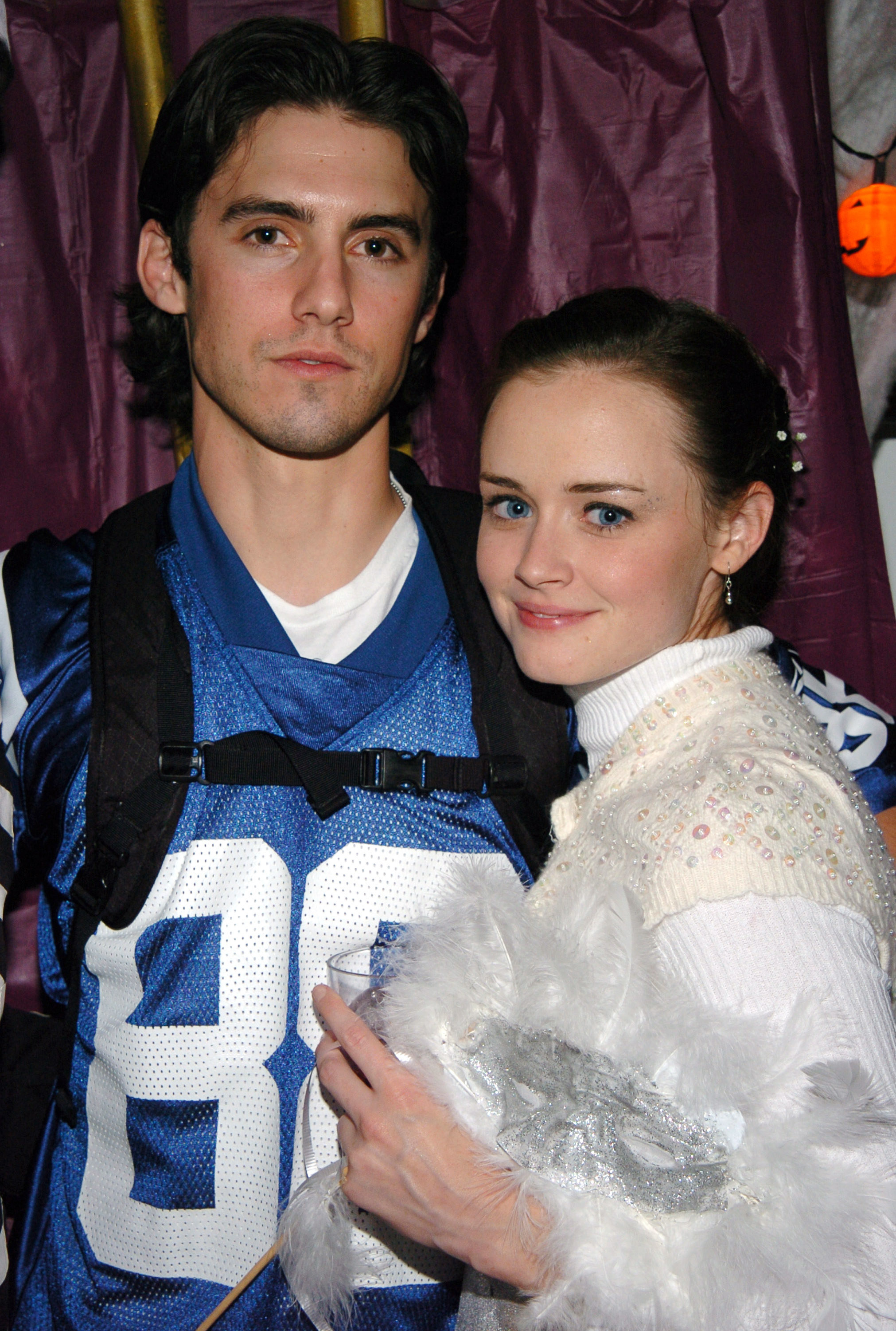 Milo Ventimiglia in a football jersey standing next to Alexis Bledel who&#x27;s wearing a  sequined sweater