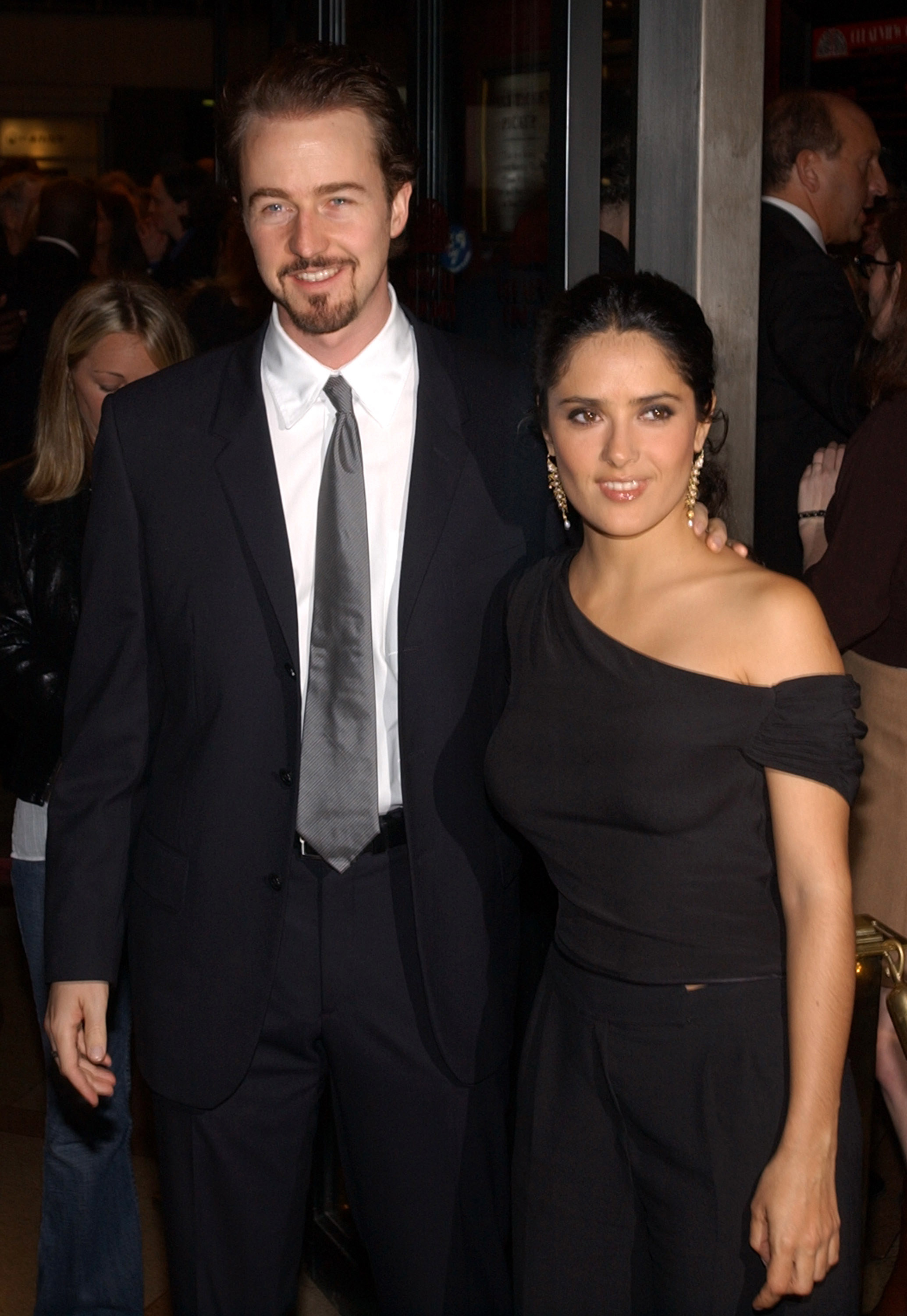 Edward Norton, wearing a suit and tie, stands with his arm across Salma Hayek&#x27;s shoulder. Salma is wearing a two-piece outfit and her hair pulled back
