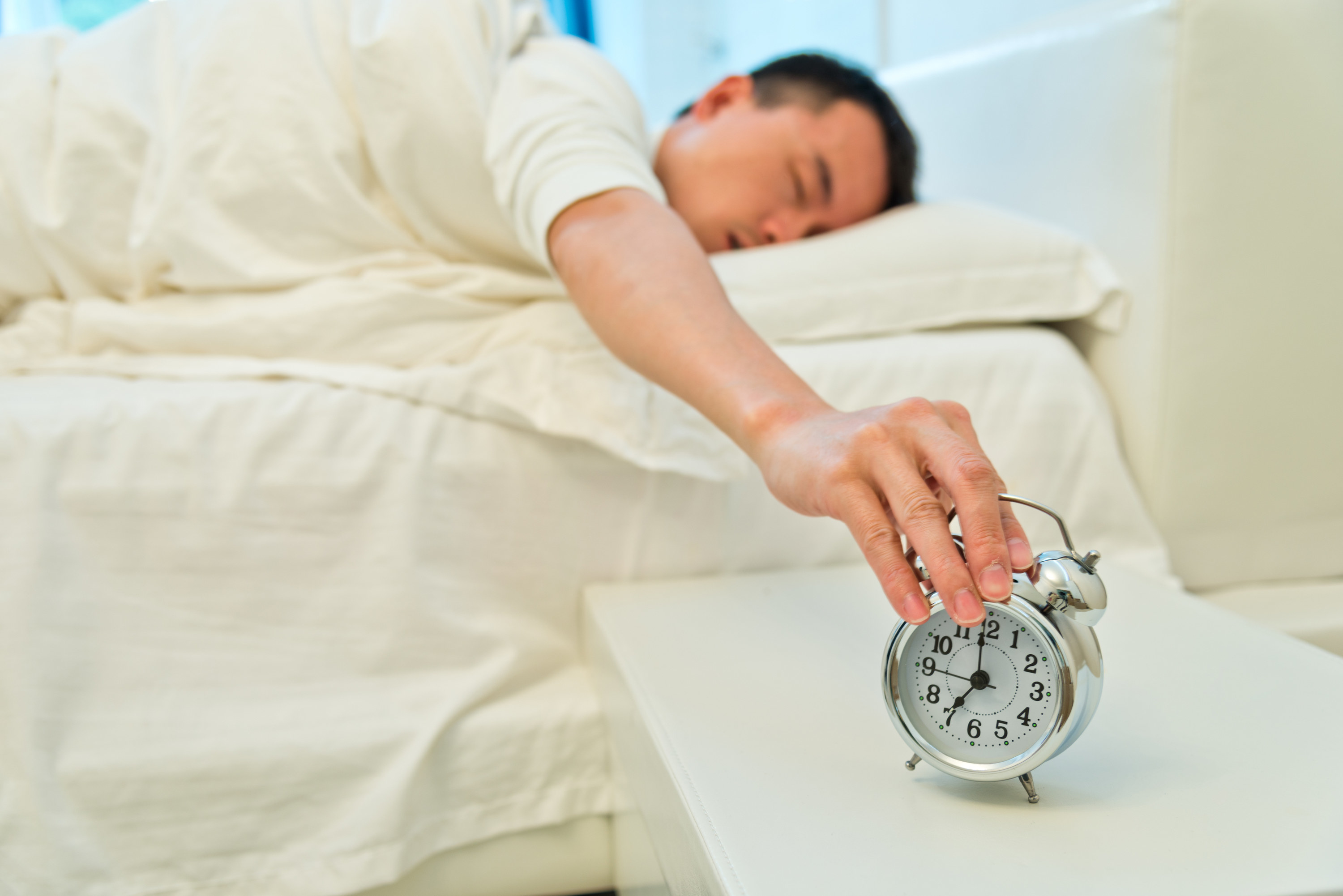 A man in bed, hitting the snooze button on his alarm clock