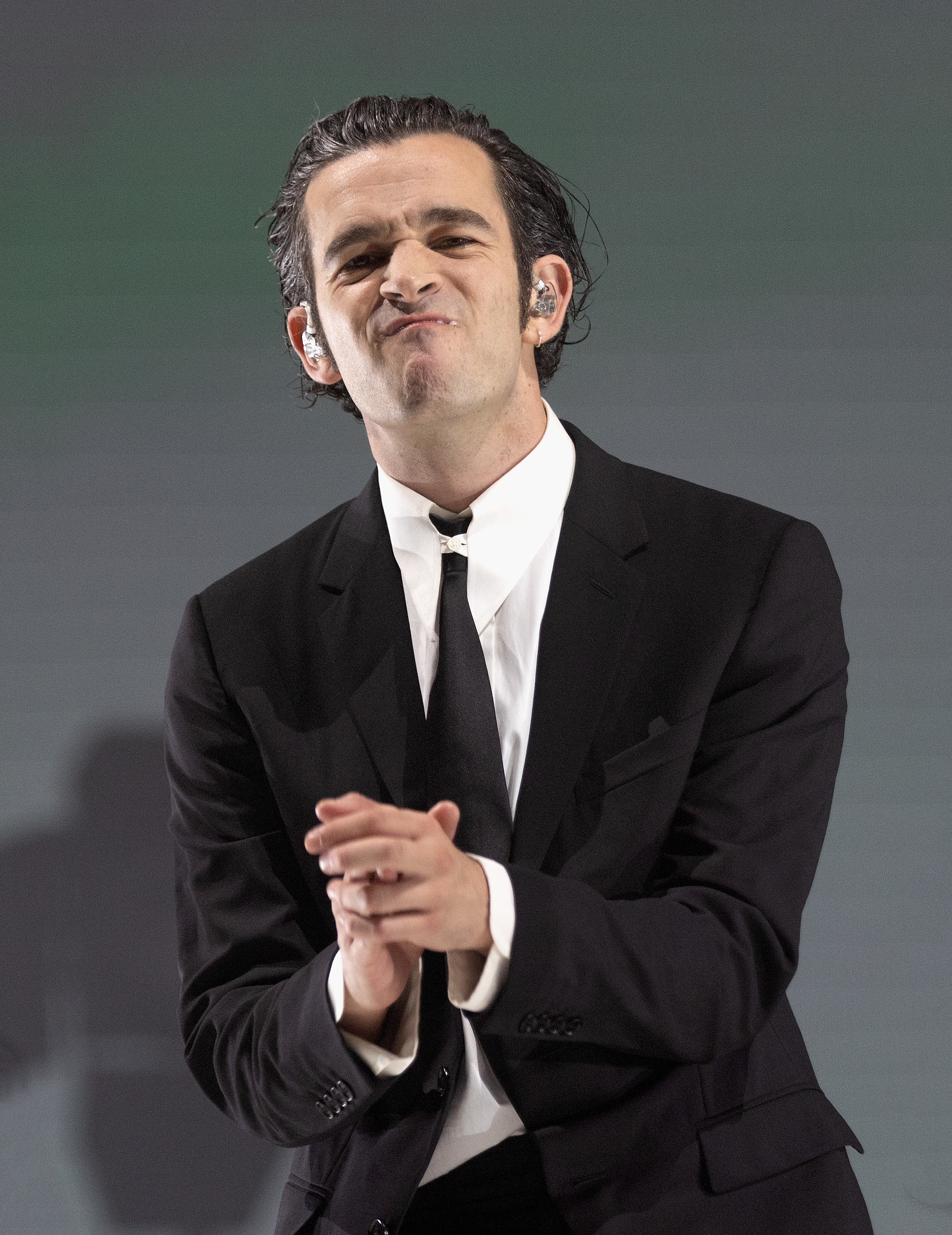 Close-up of Matty in a suit and tie and grimacing with clasped hands