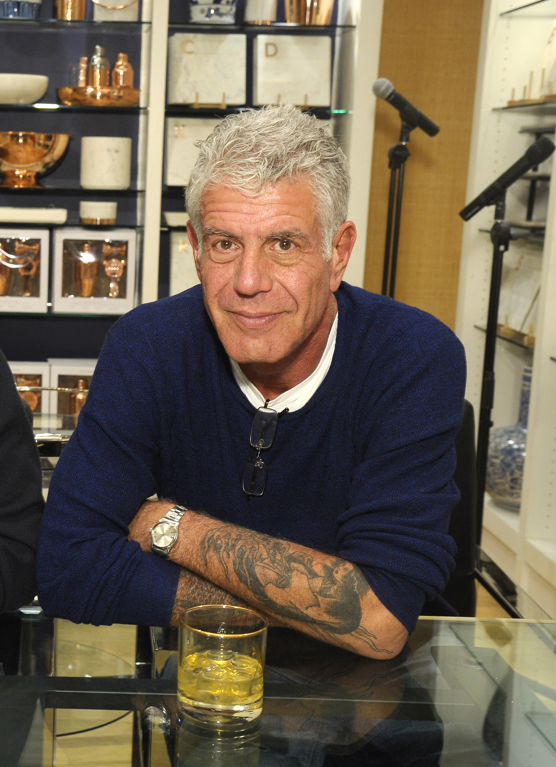 A closeup of Anthony Bourdain sitting with his arms crossed on a table with a drink