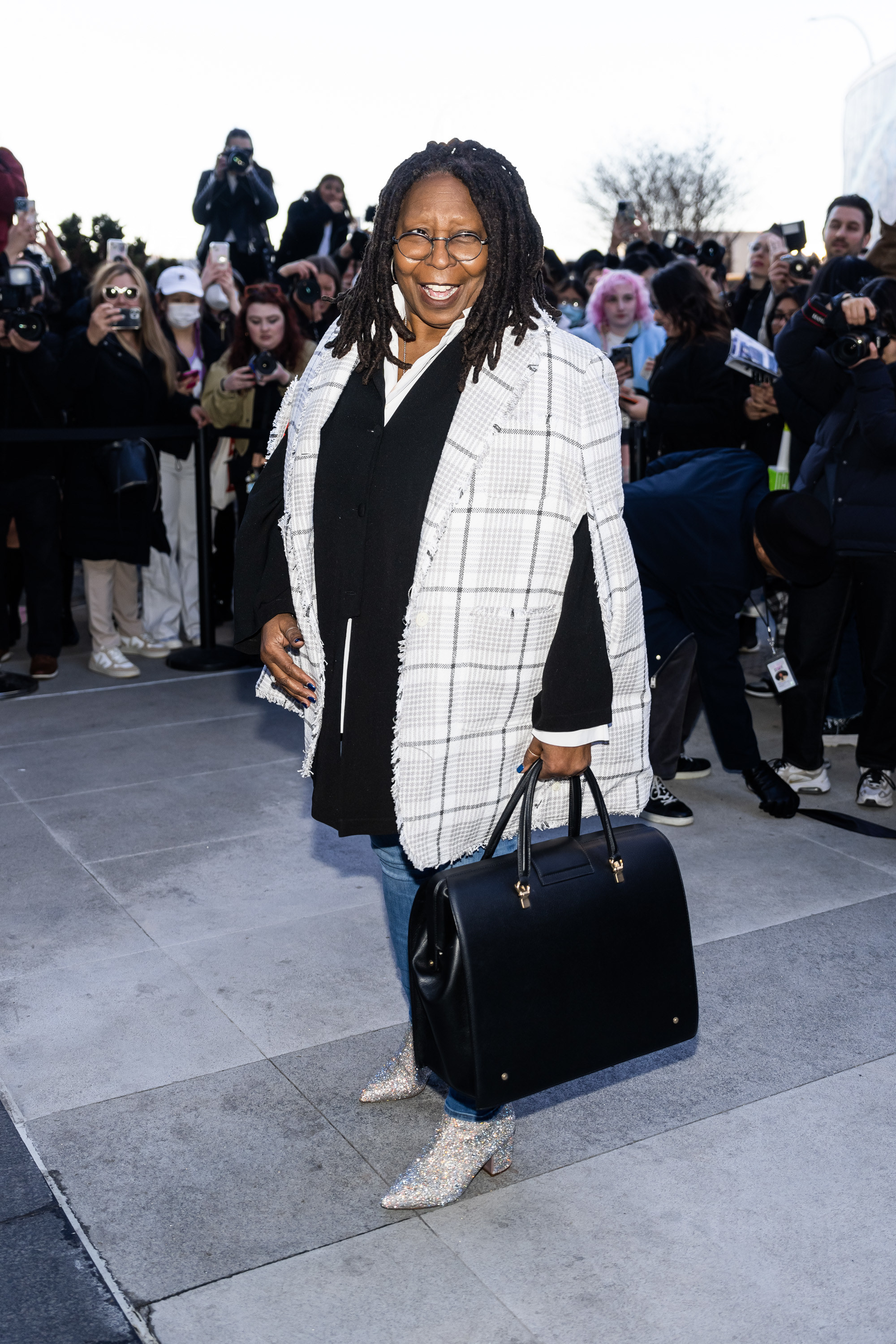 Whoopi standing outside in a plaid jacket, jeans, and sequined boots as photographers take her photo