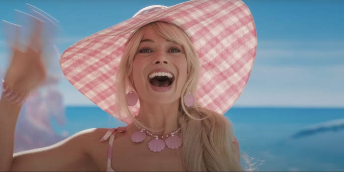 Margot Robbie as Barbie waving excitedly. Barbie is wearing a plaid pink-and-white wide-brimmed hat and a necklace with three pink seashells