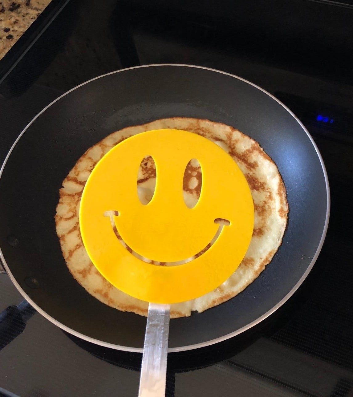 Reviewer image of the smiley face spatula on a pan with a crepe cooking in it