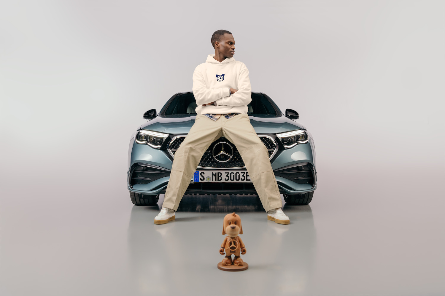 A model wears a beige sweatsuit in front of a Mercedes-Benz car next to a wooden figure made in collaboration with Superplastic.