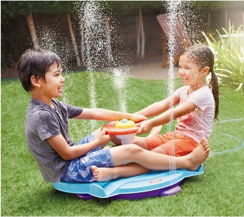 Two kids play on a sprinkler sit and spin