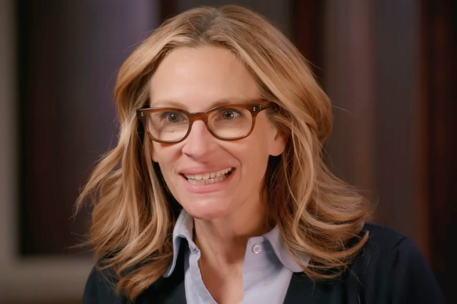 A screenshot from an interview with Julia Roberts, wearing glasses and a collared shirt, looking surprised